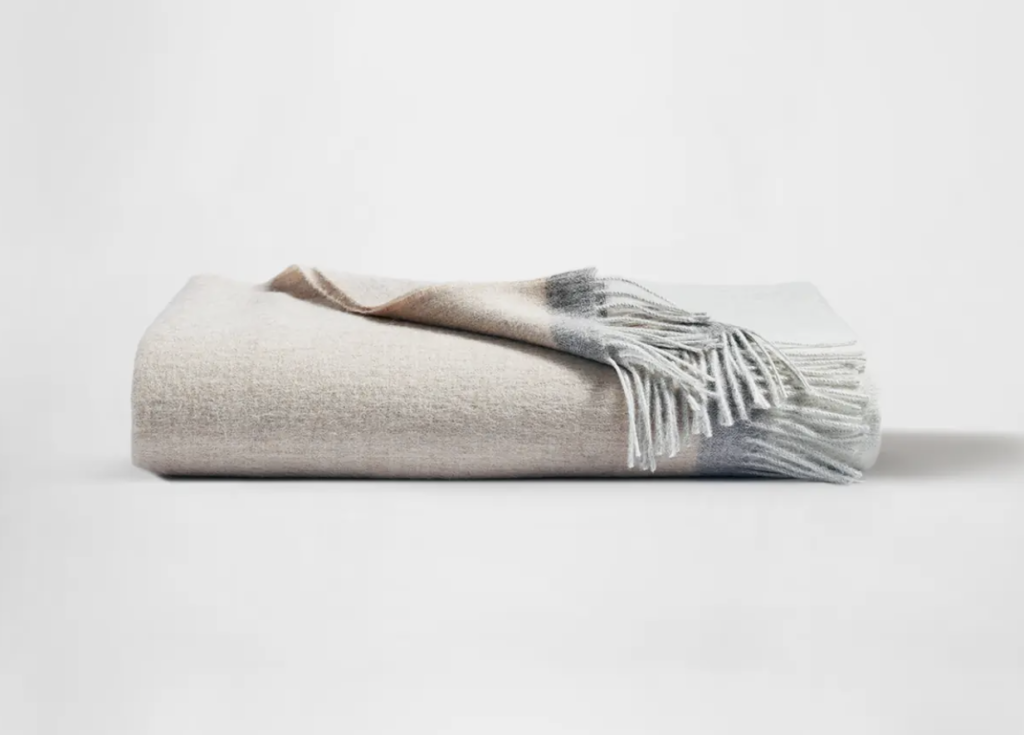 A soft, two-tone cashmere blanket in light beige and gray with fringe details.